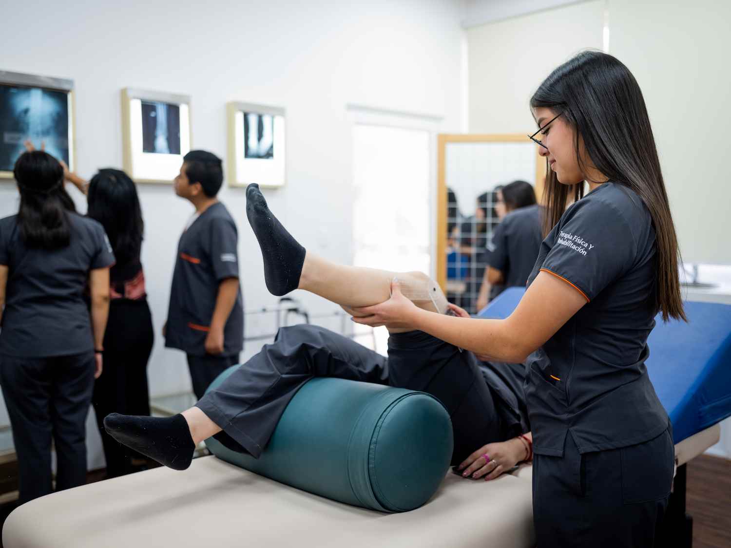 Estudiante de fisioterapia realiza movilización de pierna a paciente recostado mientras grupo analiza radiografías al fondo.